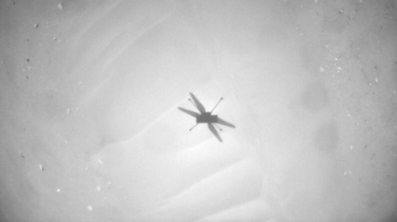 black-and-white photo showing the shadow of a small helicopter flying over sandy ground.