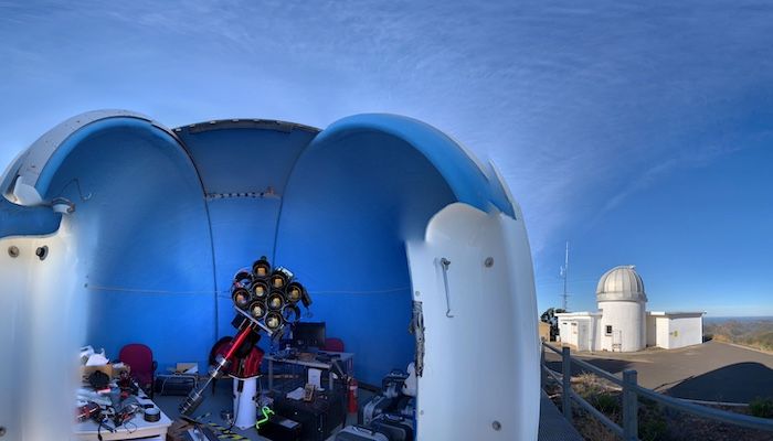 an open sided dome structure hovers above telescope equipment in the foreground. behind to the right, a domed silo with blue skies above.