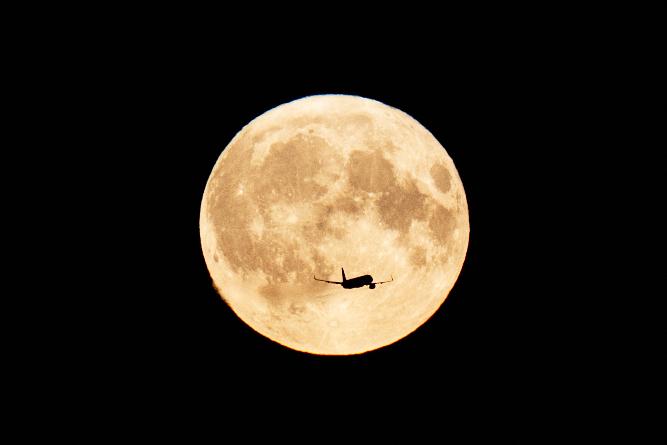 Uma lua grande e totalmente iluminada ao fundo e em primeiro plano a silhueta de um avião voando pela lua.