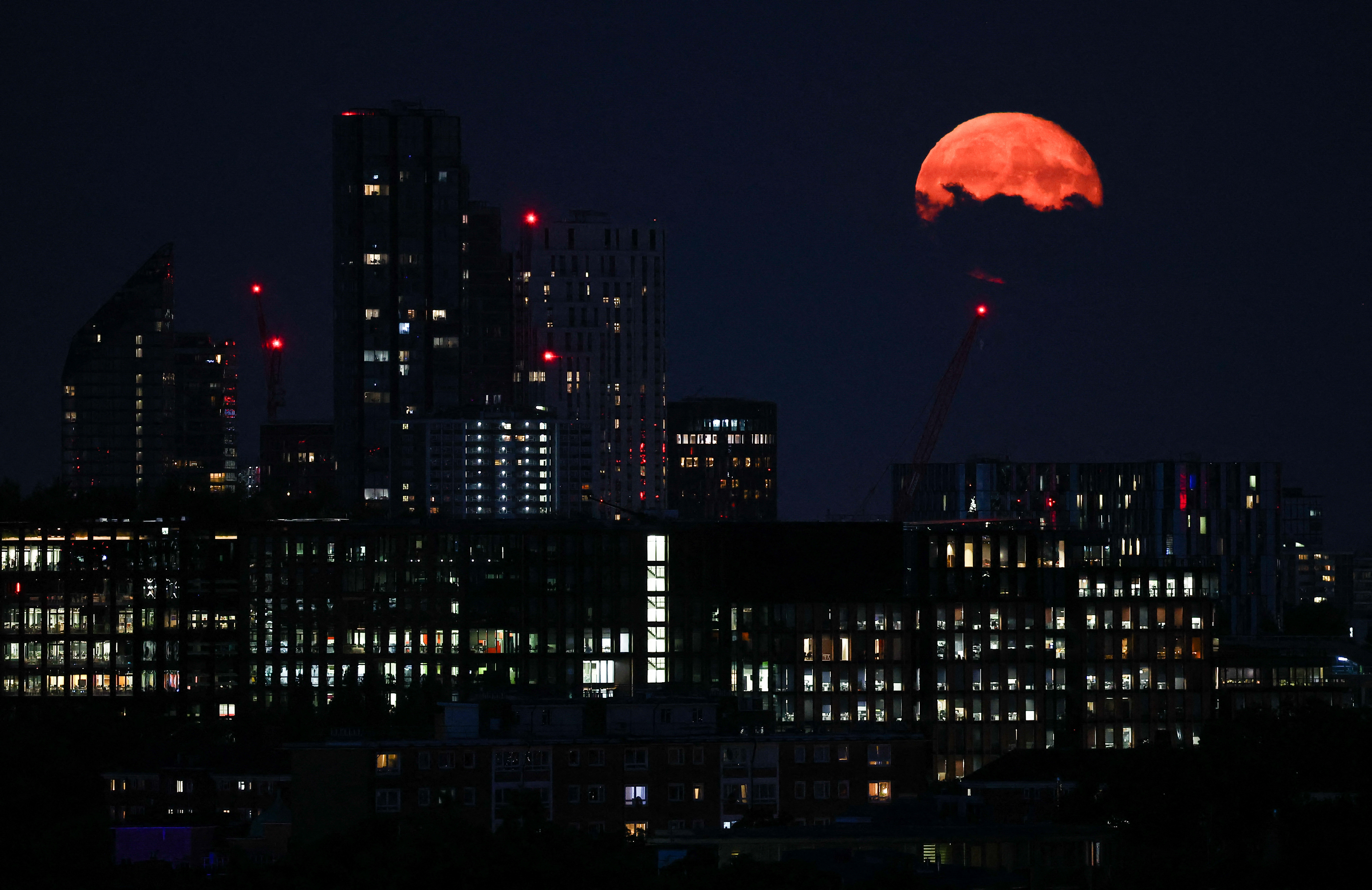 Uma cena de cidade à noite com edifícios altos em primeiro plano e uma lua brilhante em tom laranja aparecendo atrás de algumas nuvens no canto superior direito da imagem.