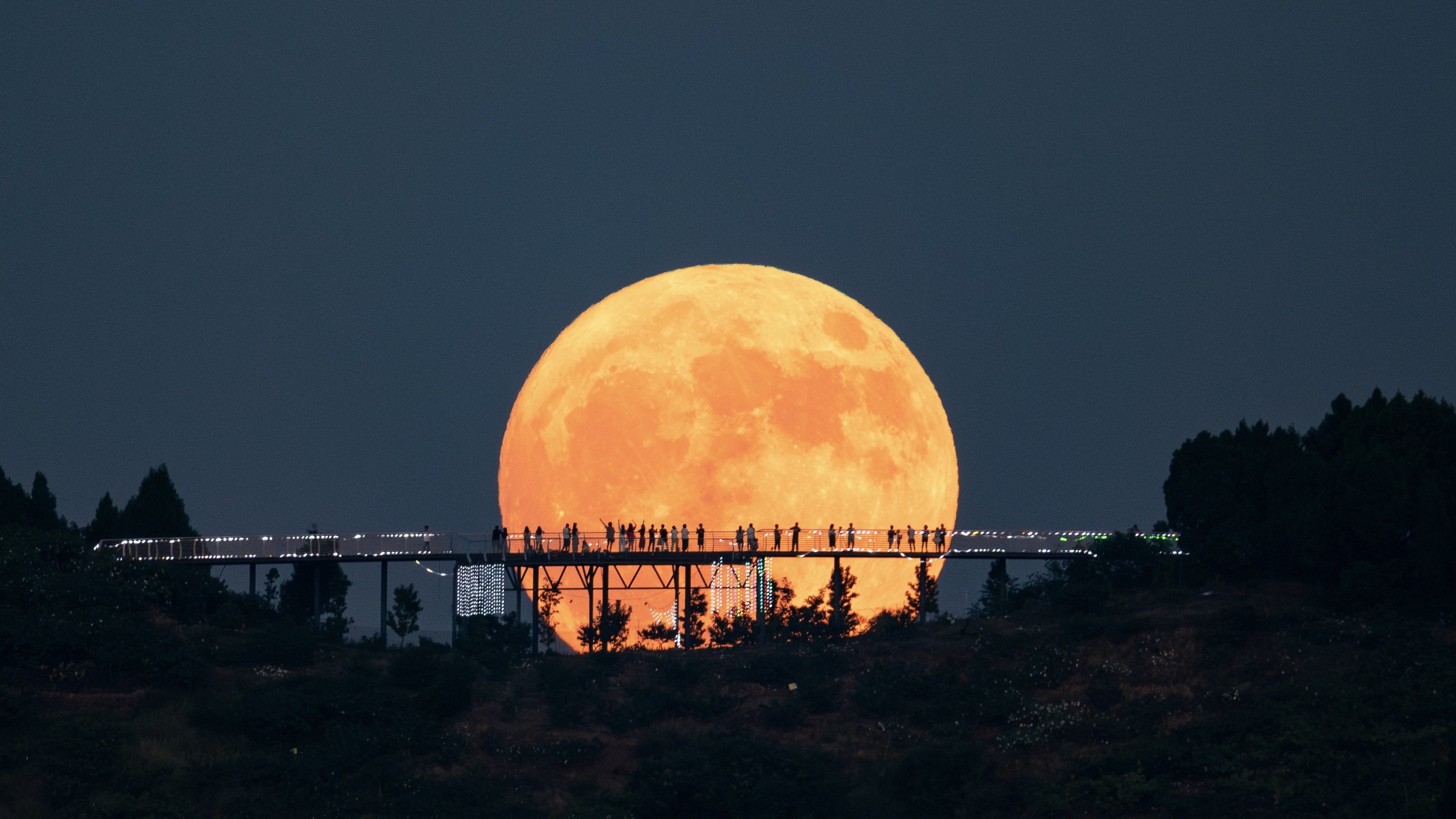 Uma grande lua brilha ao longe e, em primeiro plano, há um longo deck de observação em forma de ponte, onde as pessoas ficam de pé. Suas silhuetas se destacam em forte contraste na superfície da lua.