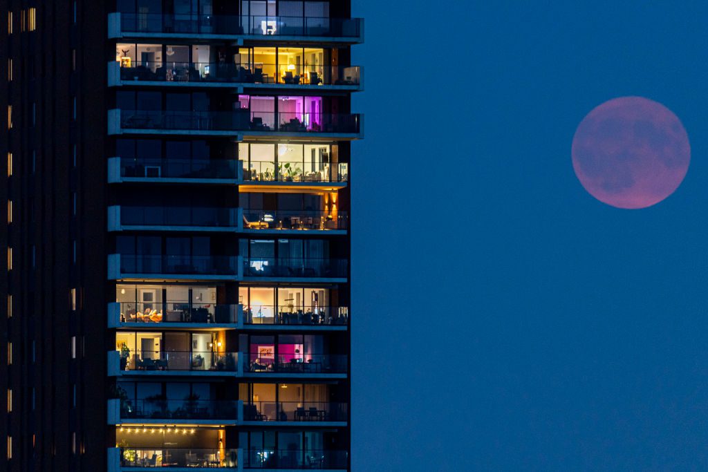 Uma lua vermelha embaçada brilha fracamente à direita de um prédio alto.